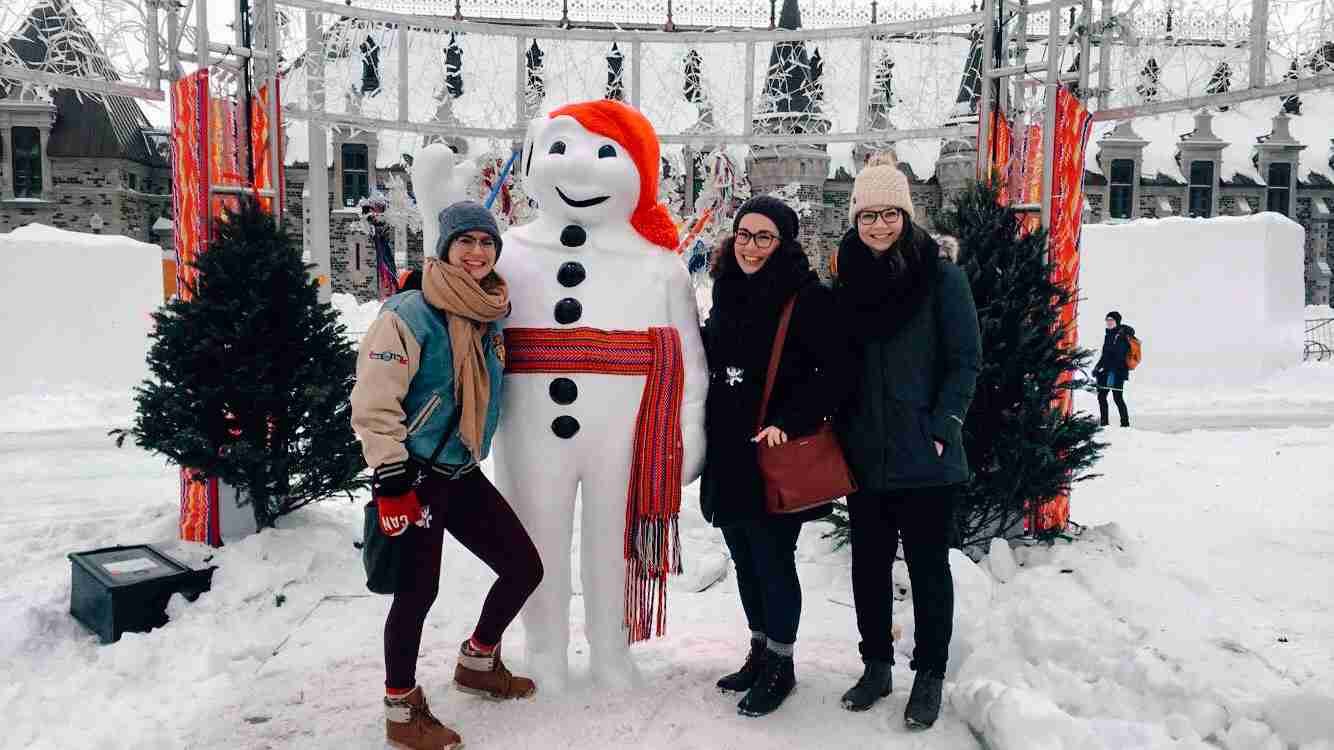 3 girls smiling with the Carnaval de Quebéc mascot- Bonhomme - with christmas trees behind them and surrounded by snow