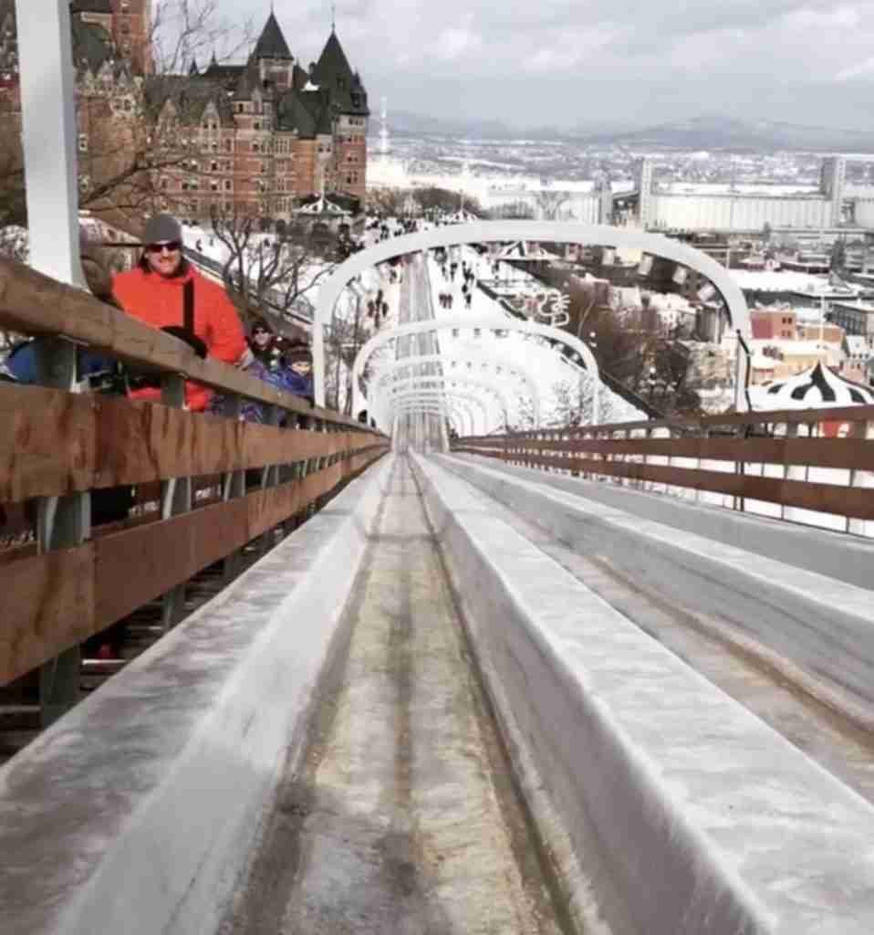 Someone sits at the top of an ice toboggan, ready to depart looking down at the track in front of them and a castle in the distance.