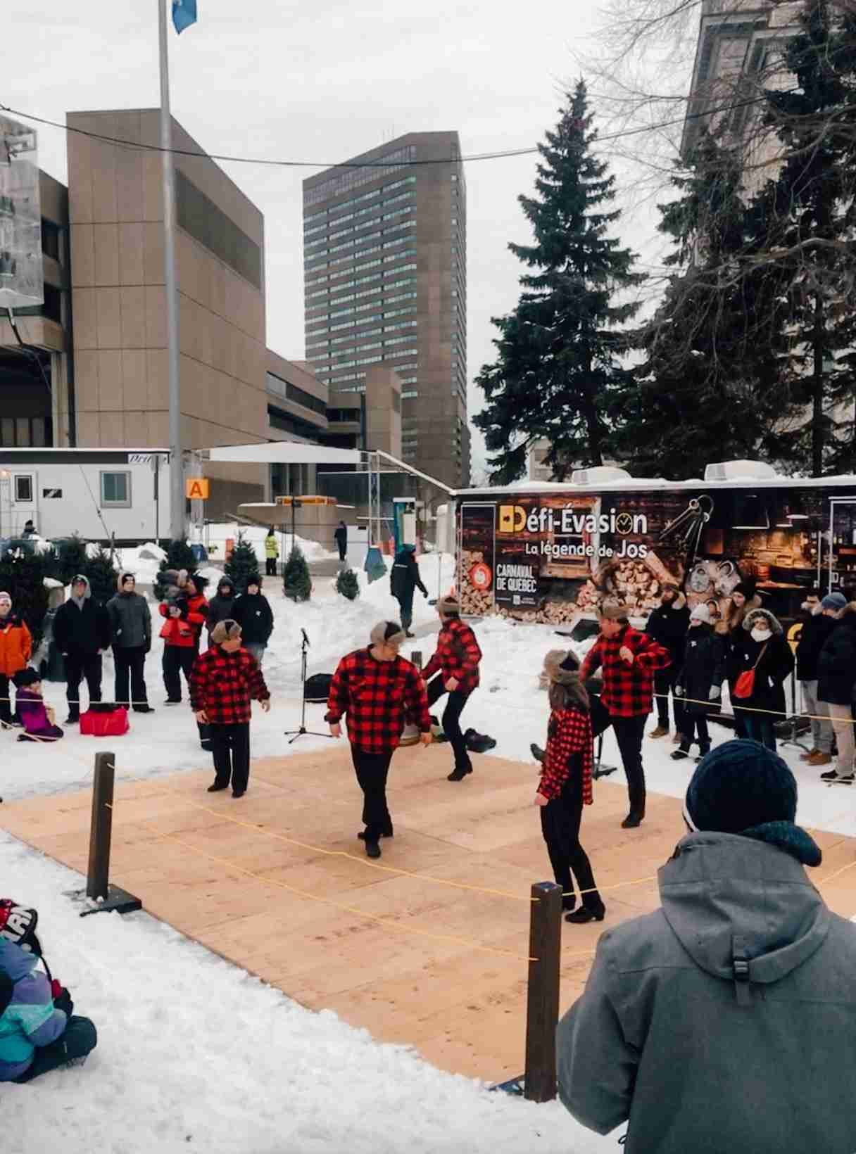 Men and women dancing on a wooden platform wearing black and red plaid while a crowd watches