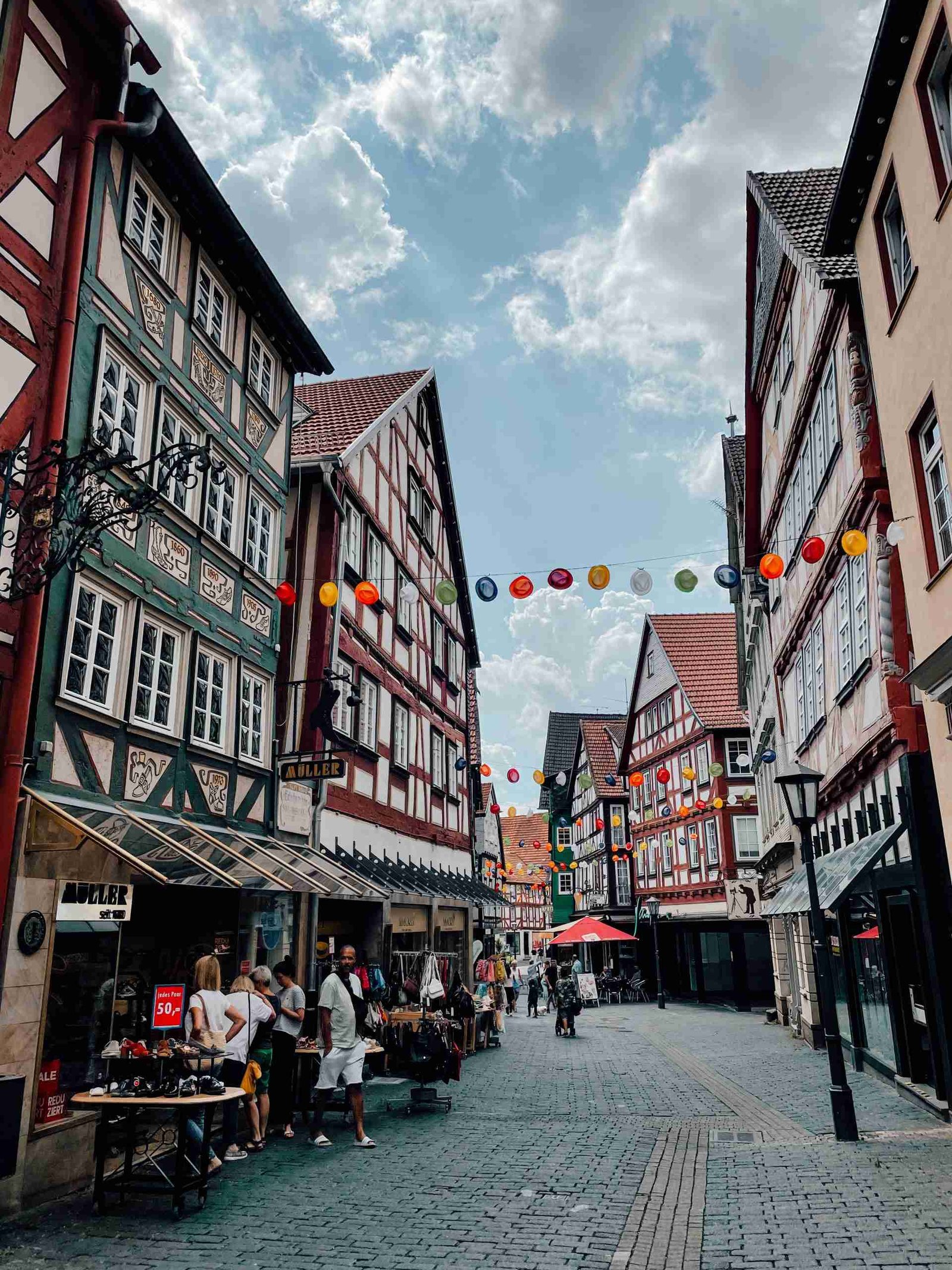 The timber-framed colorful streets of Alsfeld with a row of colorful hats hanging across the street and shoppers visiting stores.