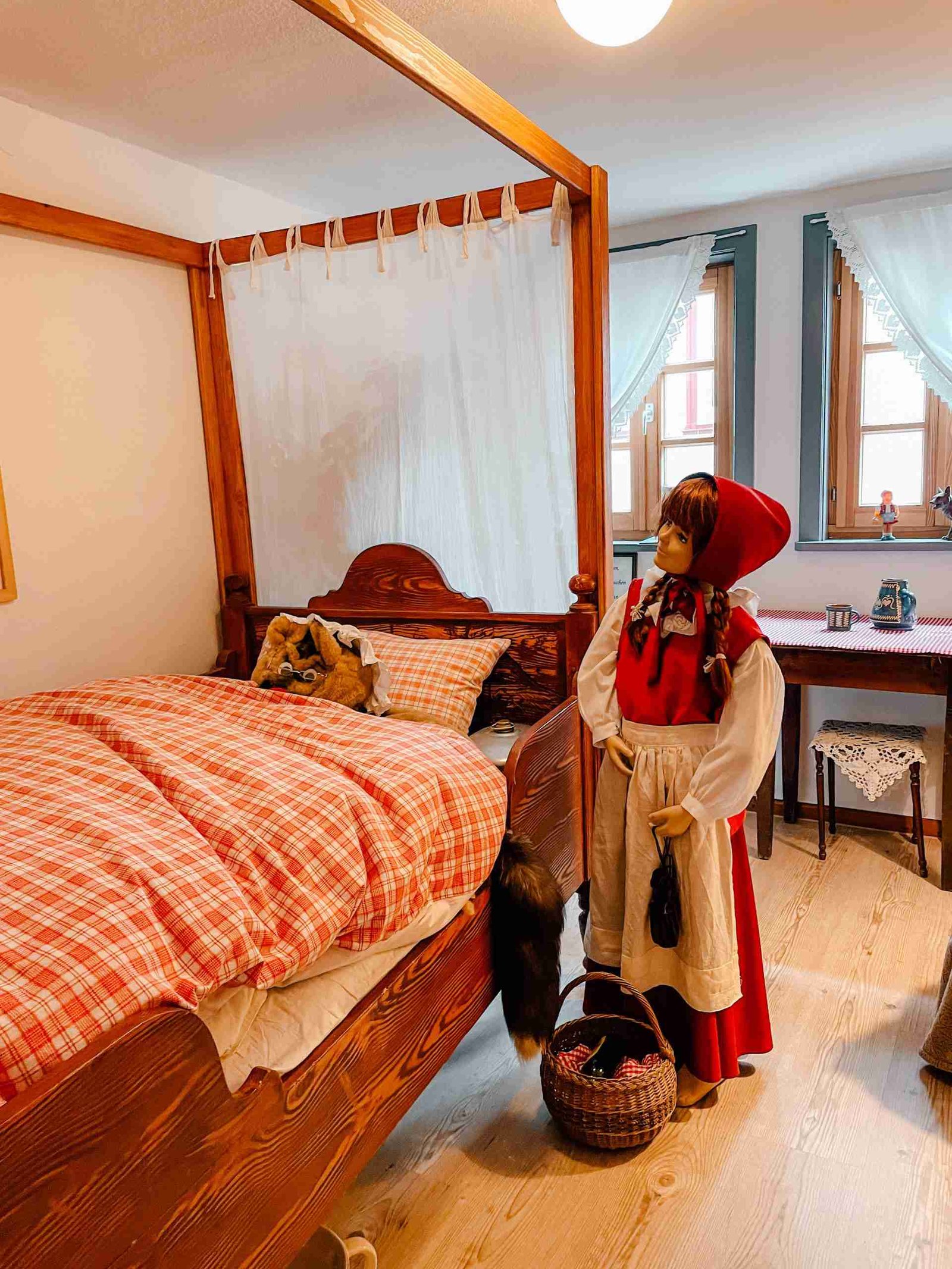 A mannequin dressed as Little Red Riding Hood standing next to a bed with a stuffed wolf under the covers - an exhibition inside Little Red Riding Hood's home in Alsfeld