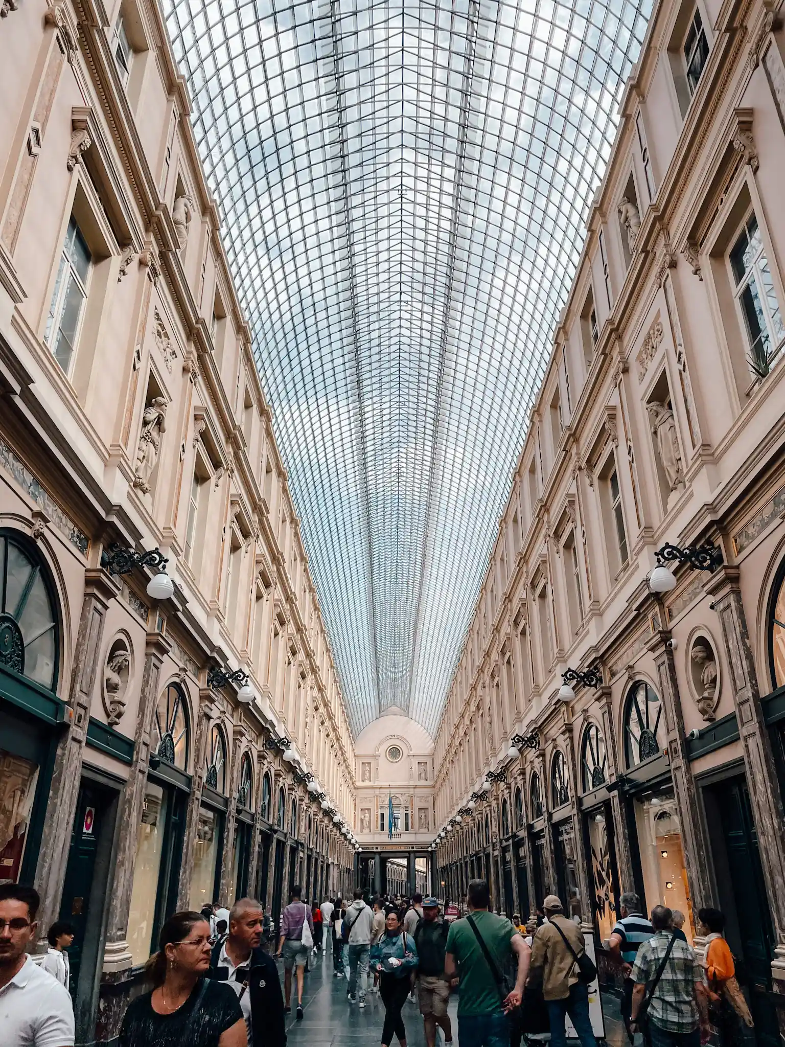 A long arcade with a glass ceiling and people crowded along the hallway. Beautiful ornate buildings line the hall