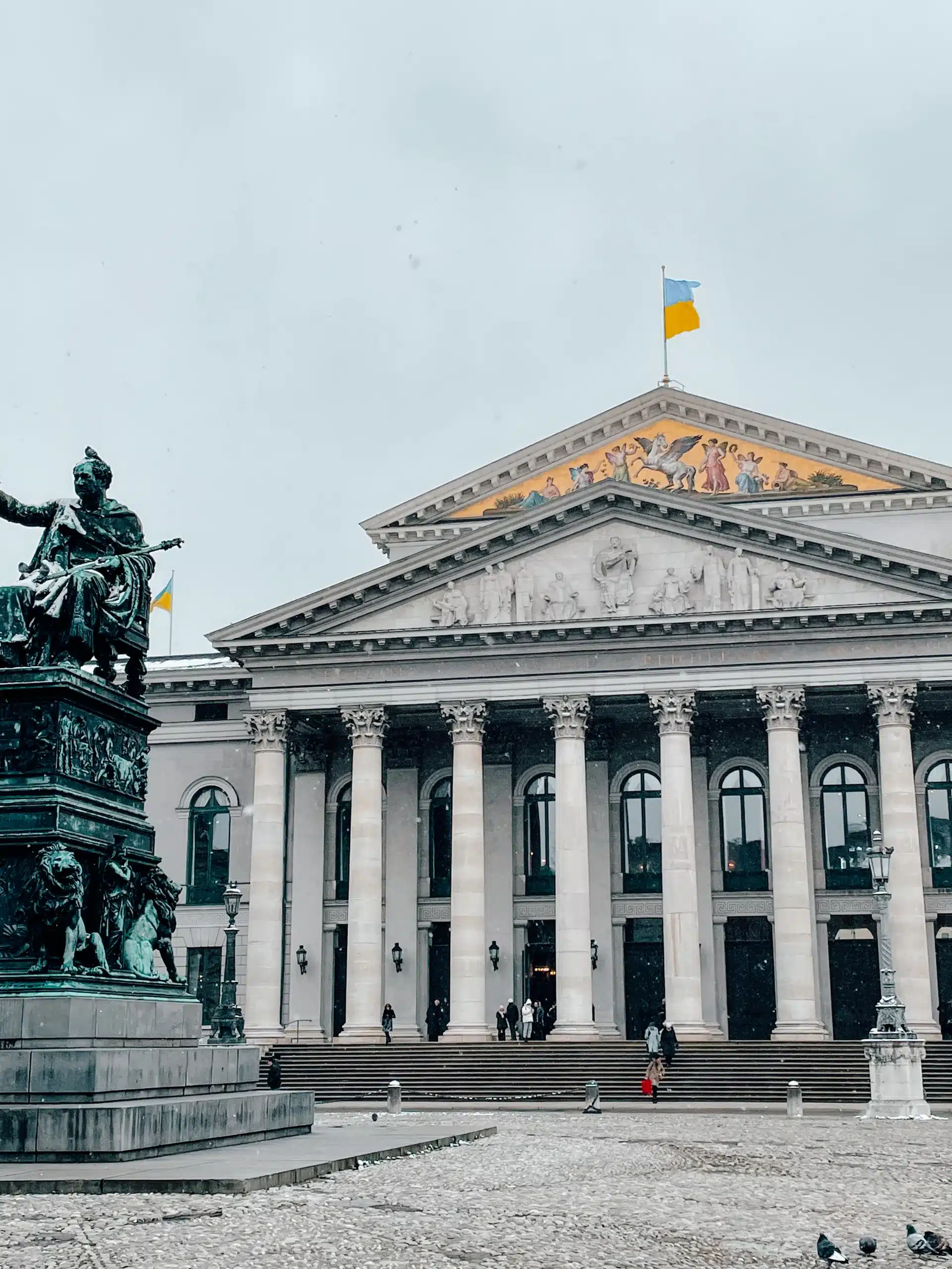 The Max Joseph Platz in Munich on a snowy day. A building stands with tall columns and a statue in front with a golden front of the Opera House! 