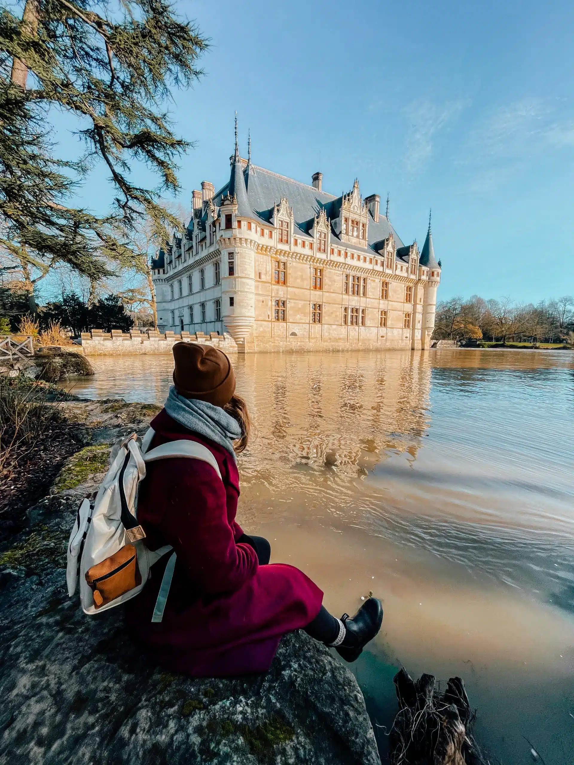 A girl sitting on a rock next to a lake looking at a white castle sitting just before her.