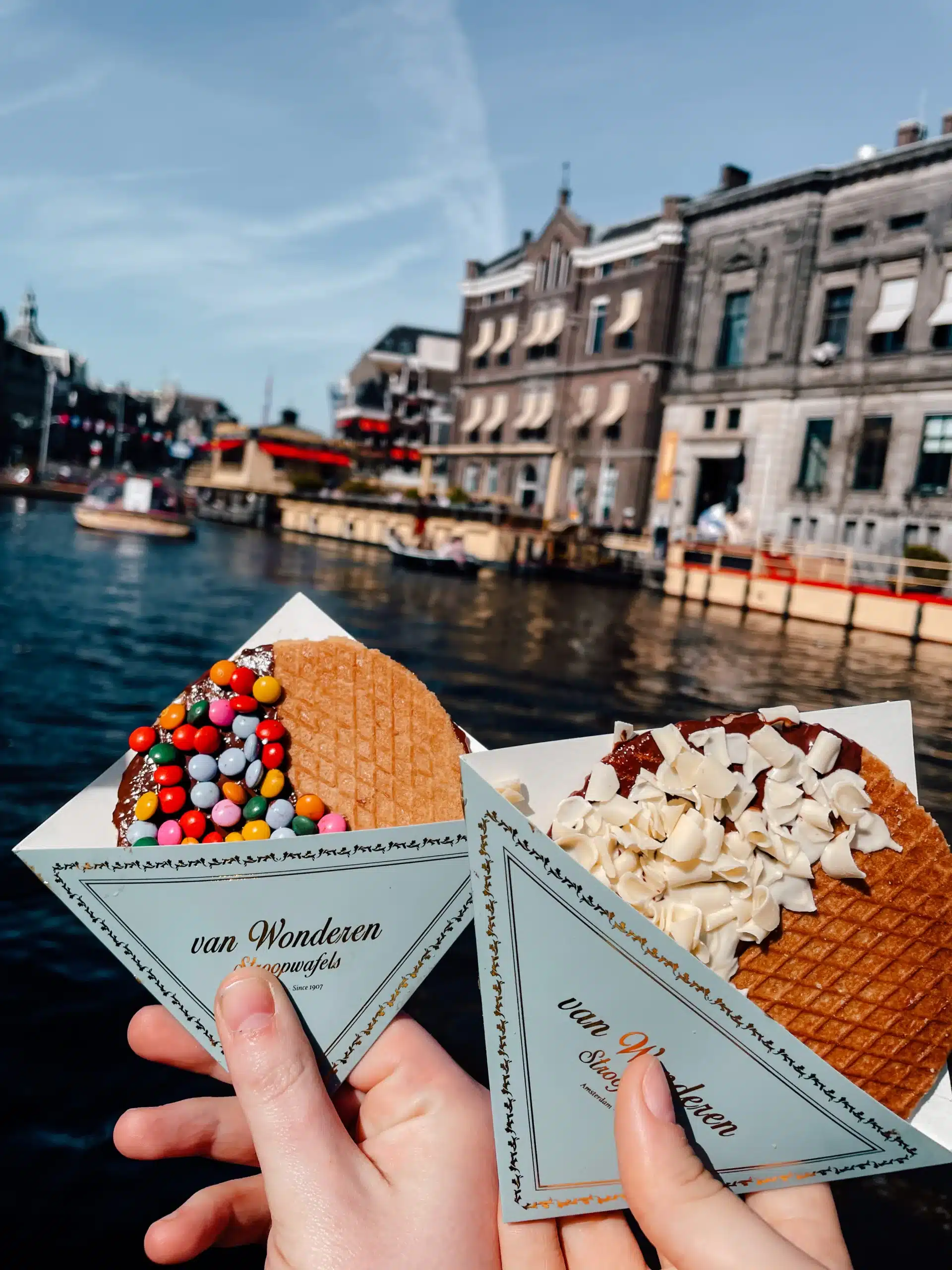 Two people holding their iconic stroopwaffels overlooking the canals, the perfect snack for an Amsterdam layover.