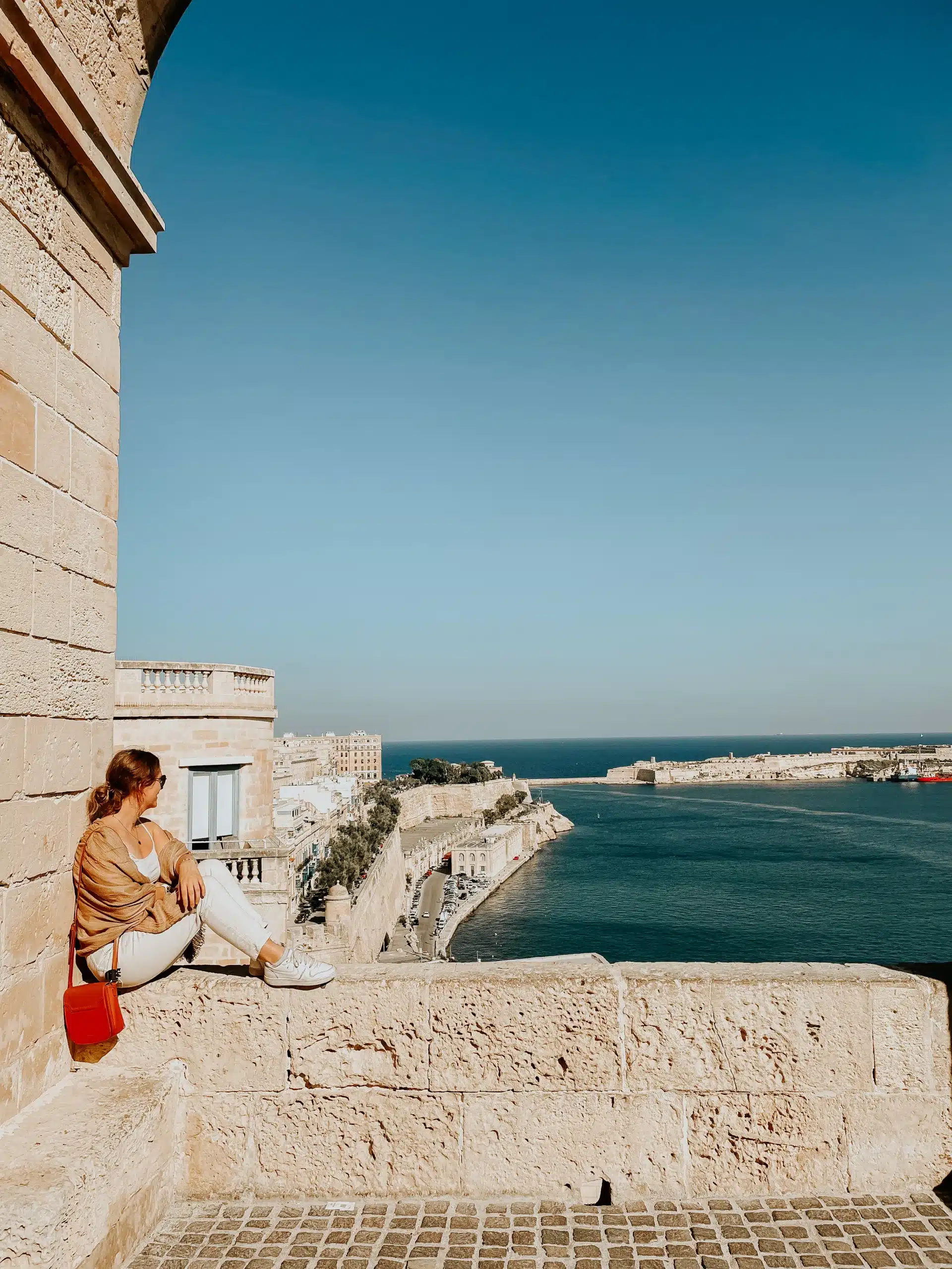 A girl sitting on a ledge overlooking the bay in Valletta on a clear, sunny day.