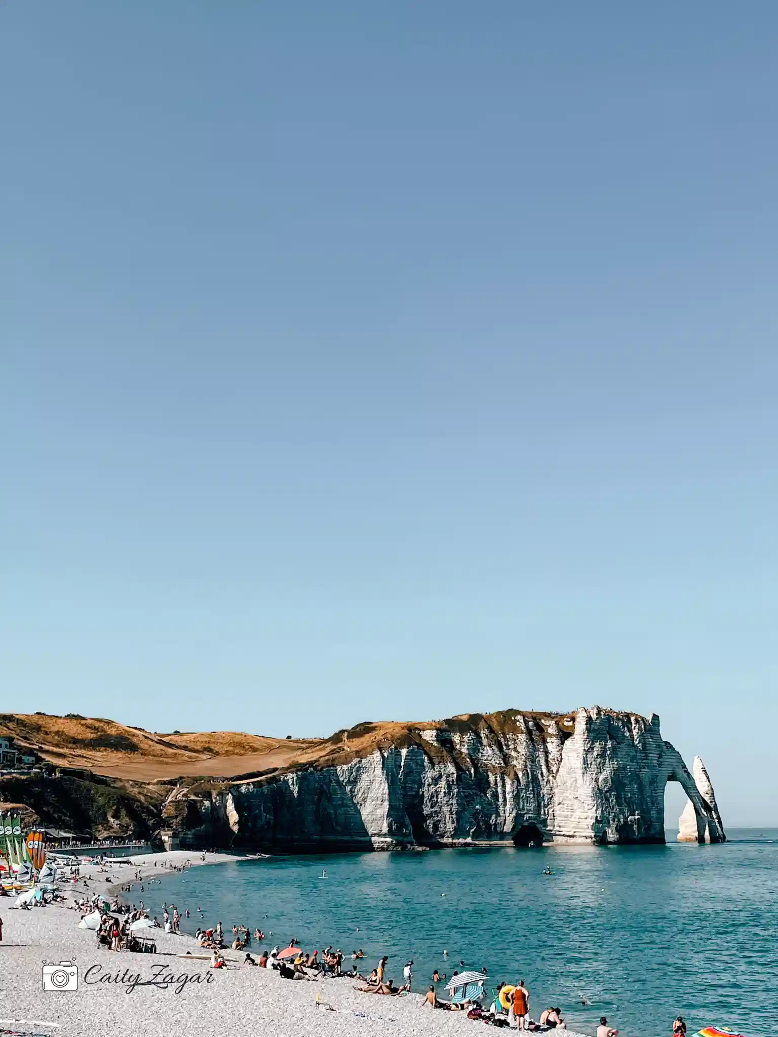 A crowd of people are enjoying the rocky beach at Etretat on a cloudless day. In front of them in the background are the white cliff formations. 