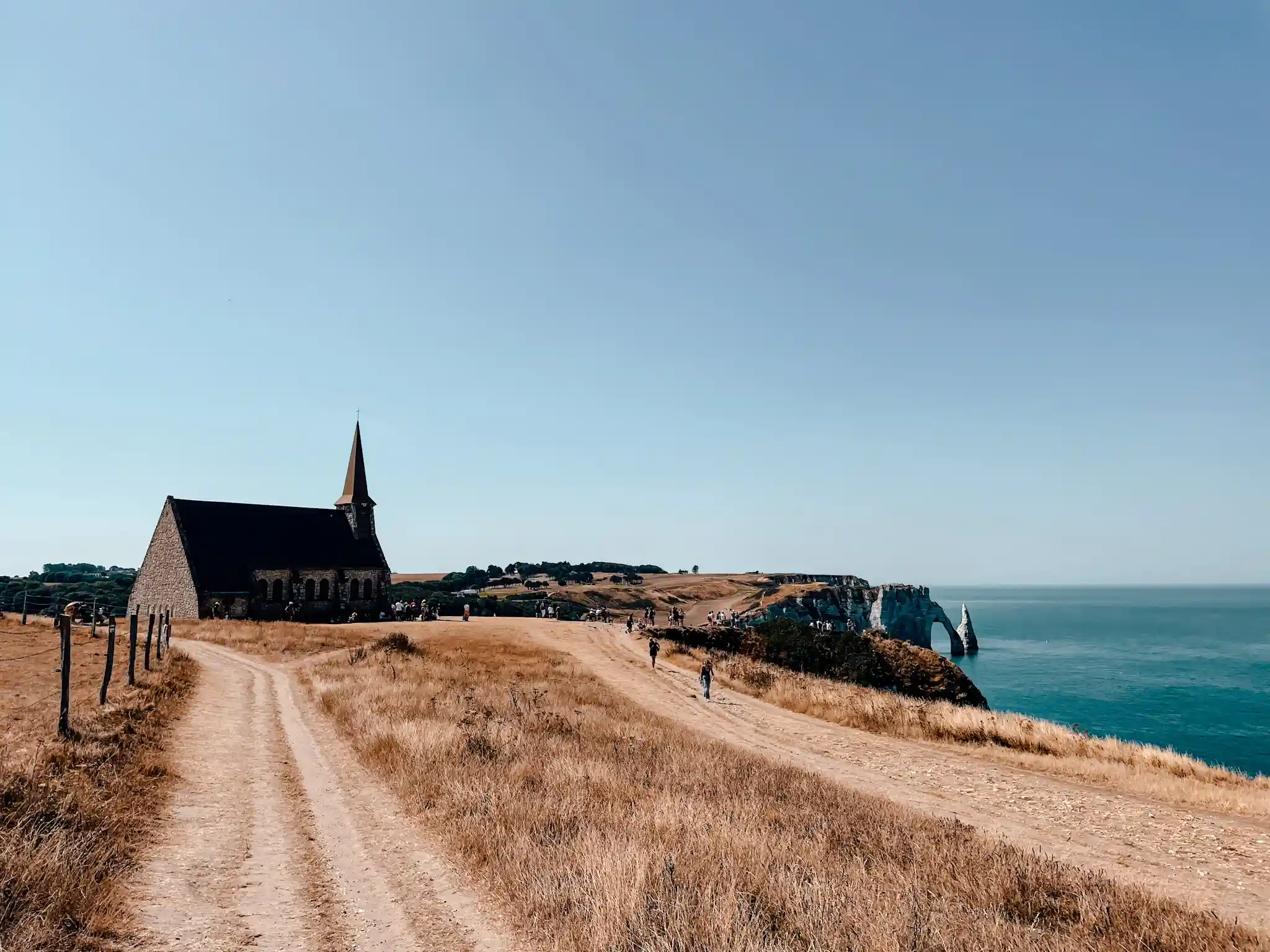 A small dark church sits a top a cliff overlooking the Alabaster coast in Etretat. People are walking along the coast enjoying the sunny day.