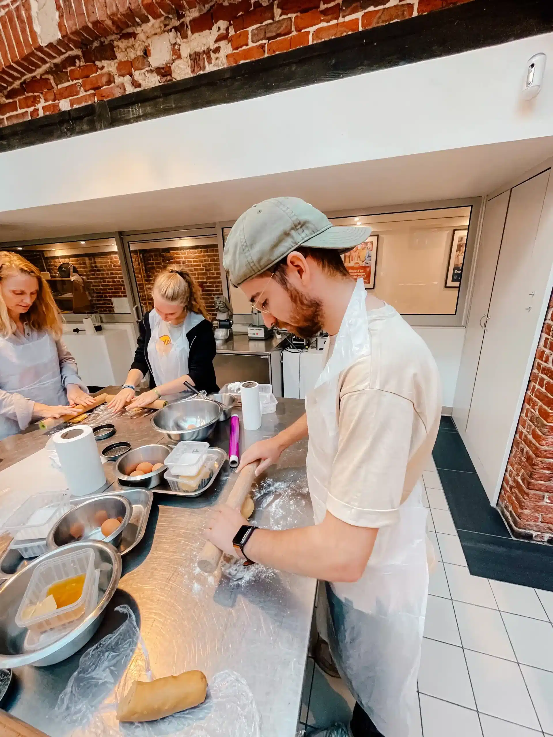 A man is standing rolling out dough in a cooking class in Paris. In front of him are different ingredients in bowls and 2 other women are working in the backgorund 