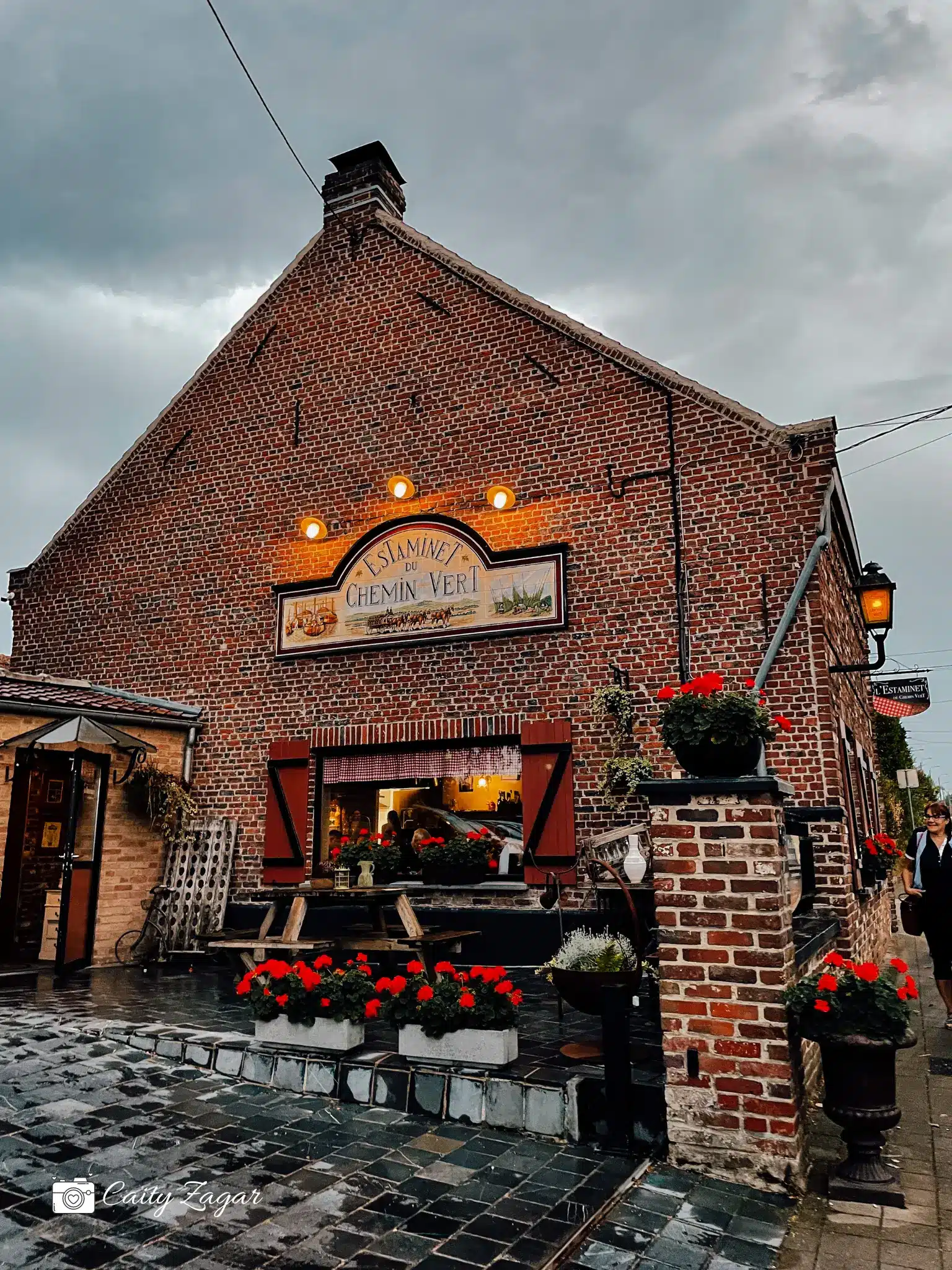 A brick building with warm outdoor lights and pots of flowers sits in front of a grey, cloudy sky in the north of France