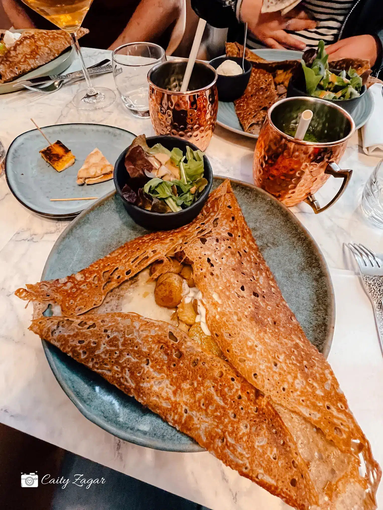 a huge, tasty galette overflowing off a circular plate with a salad and cocktail on the side 