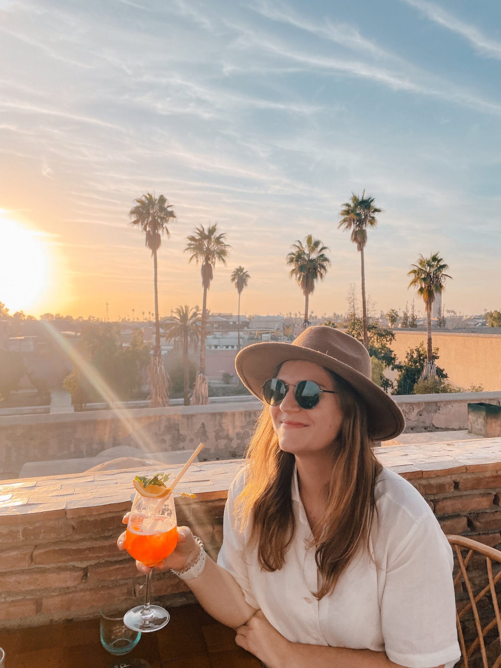 A girl sitting at a table holding a drink in Morocco while the sunsets behind tall palm trees behind her 